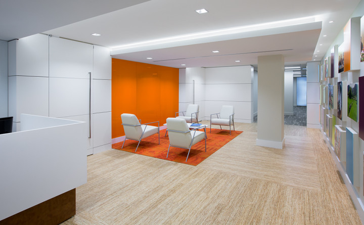 Natural wood flooring accents high-gloss white panels and back painted glass throughout the reception area. A long reception desk sits immediately to the left, across from four modern lounge chairs that sit atop a bright orange accent rug. Photo courtesy of BBGM.